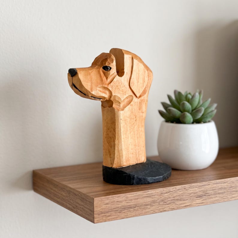 May include: A hand-carved wooden sculpture of a dog head in profile, featuring light brown wood grain, a black painted nose, and a black circular base. The dog has a smooth, stylized finish with visible carved lines defining the ears and snout.