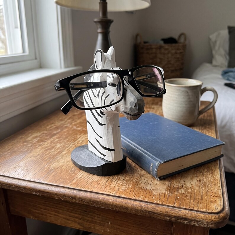May include: A hand-carved wooden zebra head eyeglass holder featuring a white base with black stripes, resting on a black oval pedestal. The zebra head is designed to support a pair of black-framed eyeglasses across its snout.