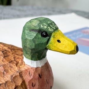 May include: A hand-carved wooden mallard duck figurine featuring a faceted texture. The head is painted green with a black eye, a white neck ring, and a bright yellow beak with black nostrils. The body displays natural wood tones with brown markings.