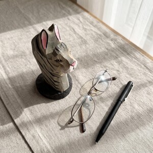May include: A hand-carved wooden cat figurine with grey and black stripes, pink inner ears, and a pink nose, positioned next to a pair of thin-framed metal eyeglasses and a sleek black ballpoint pen on a textured light grey fabric surface.