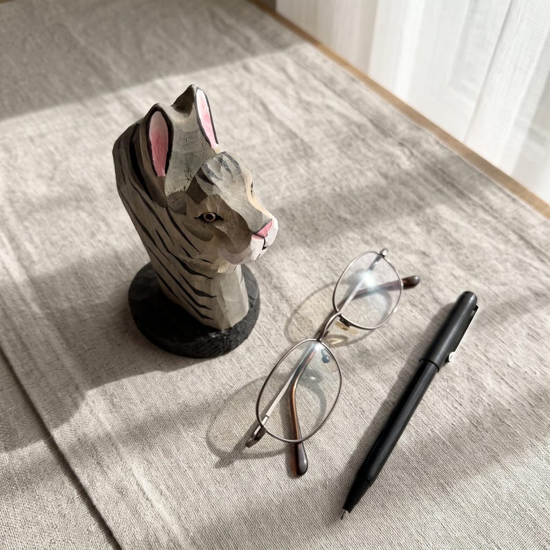 May include: A hand-carved wooden cat figurine with grey and black stripes, pink inner ears, and a pink nose, positioned next to a pair of thin-framed metal eyeglasses and a sleek black ballpoint pen on a textured light grey fabric surface.