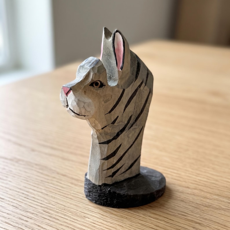 May include: A hand-carved wooden cat figurine featuring a light gray coat with dark gray stripes, a pink nose, and pink-tinted inner ears. The sculpture is mounted on a circular black base and displays visible chisel marks across its surface.
