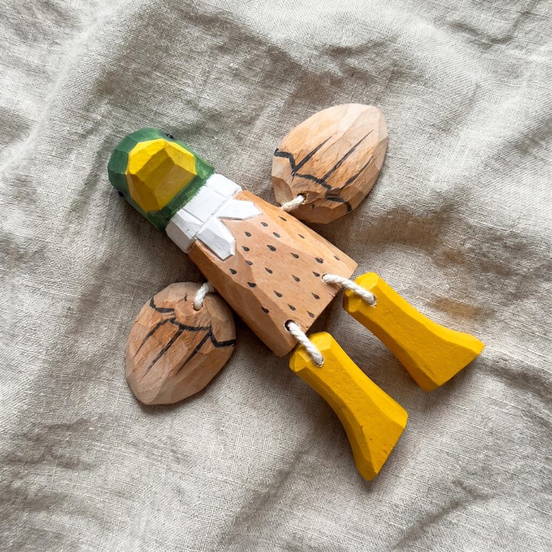 May include: A hand-carved wooden duck figurine featuring a green and yellow head, a white neck ring, and a light brown body with black painted details. The wings and yellow feet are attached to the main body with white cord, allowing for movement.