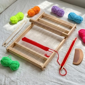 Puede incluir: Un telar de madera con una sección roja parcialmente tejida. Bolas de hilo de colores en verde, naranja, morado, azul y rosa rodean el telar. También hay herramientas de madera adicionales, como un peine y una lanzadera.