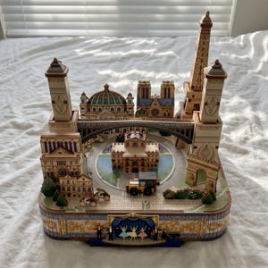 May include: A detailed decorative piece featuring Parisian landmarks such as the Eiffel Tower, Notre Dame Cathedral, and Arc de Triomphe. The base includes a stage with ballet dancers. The piece is mainly beige, with brown, blue, and gold accents.
