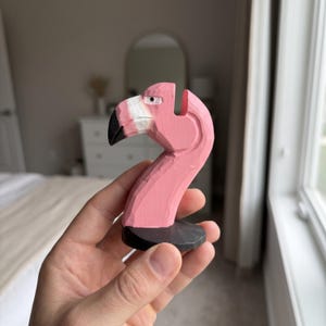 May include: A hand-carved wooden flamingo figurine painted in a vibrant pink hue. The bird features a white section near the beak, a black beak tip, and a small black eye. It is mounted on a circular black base with visible carved textures.