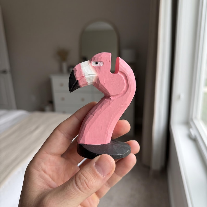 May include: A hand-carved wooden flamingo figurine painted in a vibrant pink hue. The bird features a white section near the beak, a black beak tip, and a small black eye. It is mounted on a circular black base with visible carved textures.