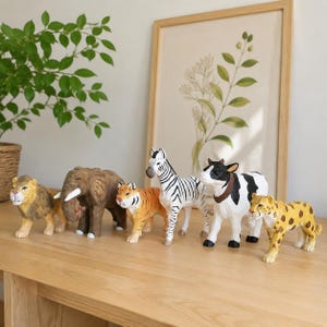 May include: A collection of wooden animal figurines, including a lion, elephant, tiger, zebra, cow, and cheetah, are arranged on a light-colored wooden surface. The figurines are painted with realistic details and colors, showcasing a safari theme.
