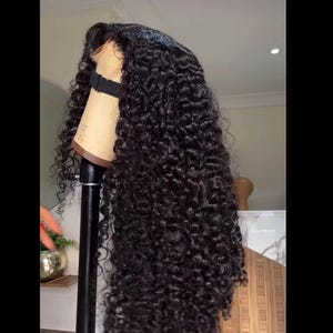 May include: A long, black curly wig displayed on a mannequin head. The wig has tight, voluminous curls and a natural hairline. The mannequin head is attached to a black stand, highlighting the wig's length and texture.