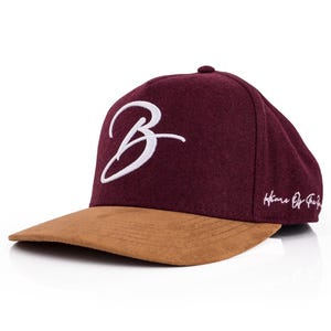 Puede incluir: Gorra de béisbol granate con visera de ante marrón. La gorra presenta una "B" blanca bordada en cursiva en la parte delantera y el texto "Home Of The" en el lateral. Un accesorio de moda.