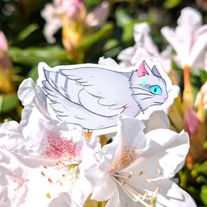 Könnte beinhalten: Ein Aufkleber mit einer stilisierten grauen Katze mit blauen Augen, rosa Ohren und einer rosa Nase. Die Katze sitzt auf einer weißen Blume mit rosa und braunen Akzenten, umgeben von grünem Laub und anderen weißen und rosa Blüten.