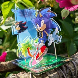 Könnte beinhalten: Ein transparenter Acryl-Aufsteller mit der blauen Figur Sonic the Hedgehog in dynamischer Pose, die rot-weiße Turnschuhe trägt. Eine kleinere Shadow-Figur und eine grüne Palme sind auf der transparenten Basis des Aufstellers abgebildet.