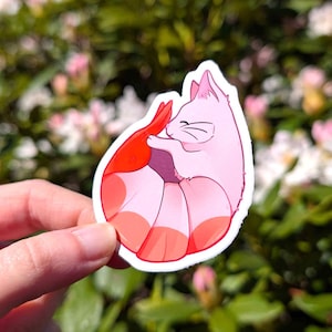 Könnte beinhalten: Ein gestanzter Aufkleber mit einer rosa Katze, die eine rote Garnele umarmt. Die Katze hat ein lächelndes Gesicht mit schwarzen Schnurrhaaren und geschlossenen Augen. Die Garnele ist rot und gebogen. Der Aufkleber hat einen weißen Rand und wird vor einem verschwommenen grünen und rosa Hintergrund gehalten.