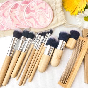 Könnte beinhalten: Eine Sammlung von Make-up-Pinseln mit hellen Holzgriffen und dunklen Borsten, zusammen mit wiederverwendbaren rosa und weißen Make-up-Entferner-Pads. Ein Bambuskamm und eine Box mit dem Text "Bamboo Comb 100% Biodegradable" sind ebenfalls enthalten.