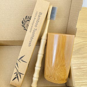 Könnte beinhalten: Ein Bambus-Zahnbürsten-Set mit einer Zahnbürste mit grauen Borsten, einem Bambusbecher und einer Pappschachtel. Auf der Schachtel und der Zahnbürstenverpackung steht "Bamboo Toothbrush 100% Biodegradable". Das Set besteht aus natürlichen Materialien.