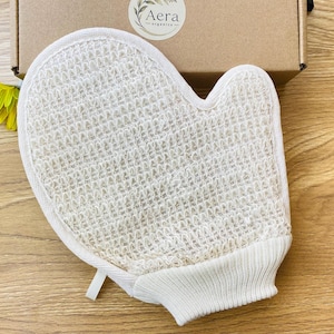 Puede incluir: Un guante de baño exfoliante de color blanco roto con un panel frontal tejido y texturizado similar a la lufa, y un puño de punto acanalado en la muñeca. El guante tiene un borde de tela clara e incluye una pequeña trabilla blanca.