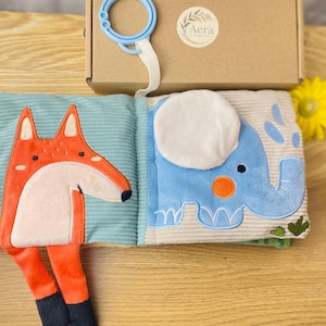 Könnte beinhalten: Offenes Stoff-Babybuch mit Fuchs- und Elefanten-Design. Der Fuchs ist orange und weiß, und der Elefant ist blau mit einem orangefarbenen Ohr. Das Buch liegt auf einer Holzoberfläche, mit einem Karton und einer gelben Blume im Hintergrund.