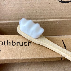 Könnte beinhalten: Nahaufnahme einer Bambus-Zahnbürste mit weißen Borsten. Der Griff hat eine hellbraune Farbe. Die Zahnbürste befindet sich in einer Pappschachtel mit dem Aufdruck "Toothbrush".