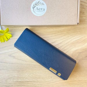 Può includere: Un astuccio rettangolare blu navy con superficie testurizzata e un piccolo dettaglio in metallo dorato nell'angolo in basso a destra. L'astuccio è posizionato davanti a una scatola di cartone marrone con un'etichetta bianca circolare con il testo Aera organics.