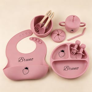 May include: A pink baby feeding set featuring a bib, bowl, plate, cup, and utensils. The set includes a bib with the name "Bruno" and a strawberry graphic. The plate also has the name "Bruno" and a strawberry graphic. The set is made of silicone.