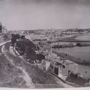 May include: Vintage black and white photograph of a coastal town. The image shows a road, buildings, and a harbor with boats. A castle-like structure is visible on a hill. The photograph is likely from the early 20th century.