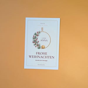 May include: A rectangular white greeting card featuring the year 2026, a circular wreath illustration with red berries and green leaves, the text LIEBE ANNA, FROHE WEIHNACHTEN, Genie&szlig;e die Feiertage!, and DEIN WICHTEL in a thin gold frame.