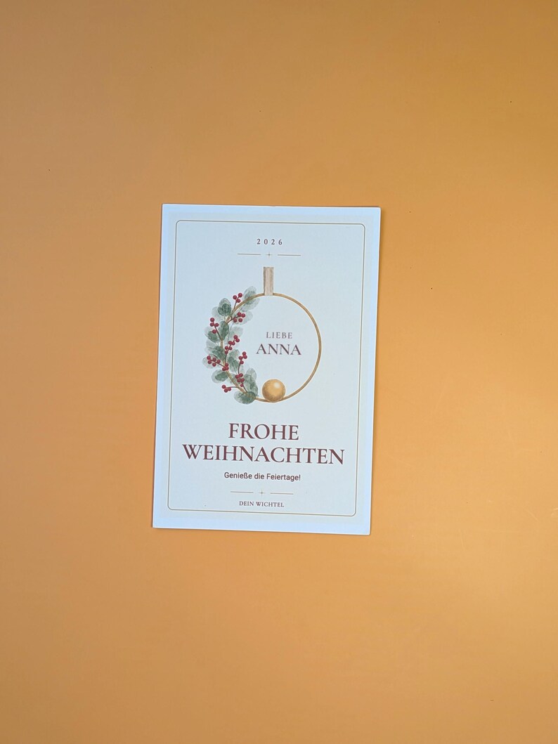 May include: A rectangular white greeting card featuring the year 2026, a circular wreath illustration with red berries and green leaves, the text LIEBE ANNA, FROHE WEIHNACHTEN, Genie&szlig;e die Feiertage!, and DEIN WICHTEL in a thin gold frame.