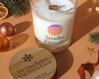 May include: A clear glass candle with white wax and a wooden wick, featuring a circular label with a sunset gradient and the text Studio Rituale. The bamboo lid is engraved with a snowflake, the text Für Oma Renate Von deinen Enkeln, and 2026.