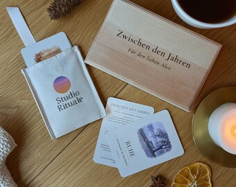 May include: A wooden box labelled "Zwischen den Jahren Für den lieben Alex" next to a white fabric pouch with a "Studio Rituale" logo. Includes cards with "RUHE" text, dried orange slices, pinecones, and a white candle in a gold holder.