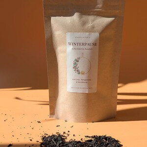 May include: A tan paper pouch of loose leaf tea with a white label featuring the text STUDIO RITUALE, WINTERPAUSE, Eine kleine Auszeit, Earl Grey, Bergamotte & Kornblume, and ZEIT ZUM DURCHATMEN, with a wreath illustration and scattered tea leaves.