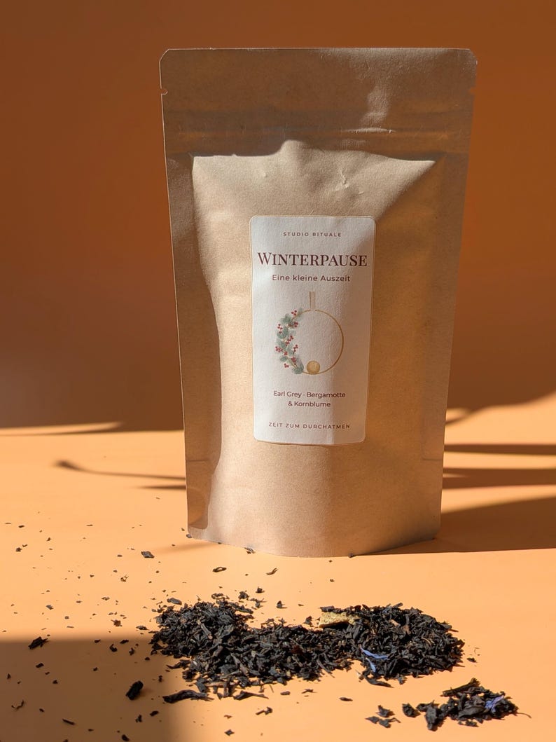 May include: A tan paper pouch of loose leaf tea with a white label featuring the text STUDIO RITUALE, WINTERPAUSE, Eine kleine Auszeit, Earl Grey, Bergamotte & Kornblume, and ZEIT ZUM DURCHATMEN, with a wreath illustration and scattered tea leaves.