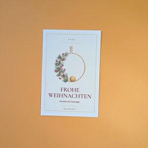 May include: A rectangular white card featuring the text 2026, FROHE WEIHNACHTEN, Genie&szlig;e die Feiertage!, and DEIN WICHTEL. The design includes a gold hoop decorated with green leaves, red berries, and a small gold sphere at the bottom centre.