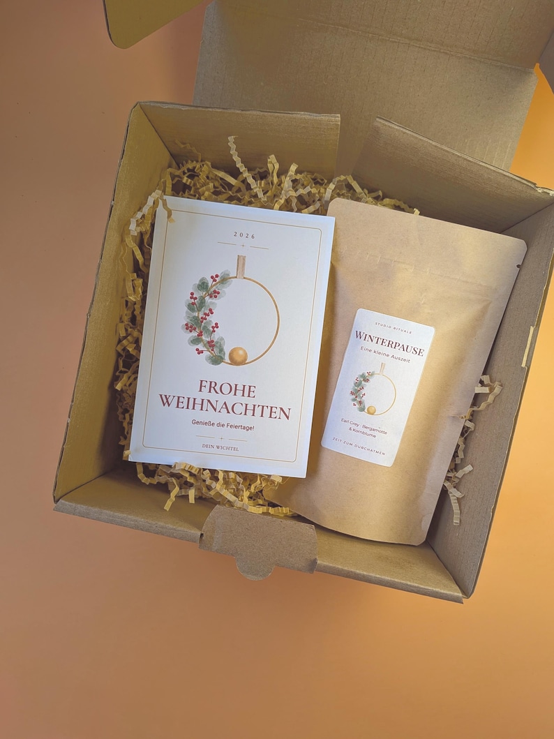 Könnte beinhalten: Ein Geschenkkarton aus Pappe mit einer weißen Grußkarte mit dem Text FROHE WEIHNACHTEN und einem Kranz-Motiv sowie einem braunen Papierbeutel mit der Aufschrift WINTERPAUSE und passender Grafik, eingebettet in Füllmaterial aus Papierstreifen.