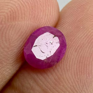 May include: A faceted, oval-shaped ruby gemstone. The gem is a deep, saturated pinkish-red color with multiple facets that reflect light. The ruby is held in a hand, showing the gem's size and shape.