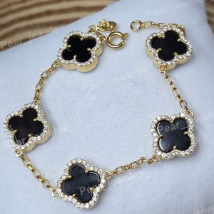 May include: Gold-toned bracelet featuring five black clover-shaped charms, each outlined with small, clear crystals. The charms are connected by a delicate chain, creating an elegant and eye-catching piece of jewelry.