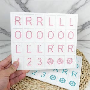 May include: White rectangular tiles with pink letters, numbers, and flower designs. The tiles are held in a hand, with a second tile visible below. The letters include R, L, and O. Numbers 1, 2, and 3 are also present.