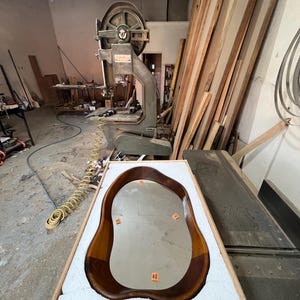 May include: An irregular, organic-shaped mirror with a polished dark brown wooden frame, resting inside a rectangular wooden crate lined with white protective foam. The mirror surface features four small orange rectangular adhesive labels.