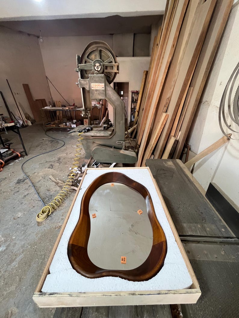 May include: An irregular, organic-shaped mirror with a polished dark brown wooden frame, resting inside a rectangular wooden crate lined with white protective foam. The mirror surface features four small orange rectangular adhesive labels.