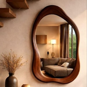 Puede incluir: Un espejo de pared con forma orgánica irregular y marco de madera oscura. El espejo refleja el interior de una sala con un sofá, una lámpara de pie y cortinas. Debajo del espejo hay un banco de madera, un jarrón con ramas secas y libros.