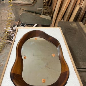 May include: An irregular, organic-shaped mirror with a polished dark brown wooden frame. The mirror surface features three small orange square stickers with the text YILDIZ CAM printed on them, and it is securely packed in a white foam-lined wooden crate.
