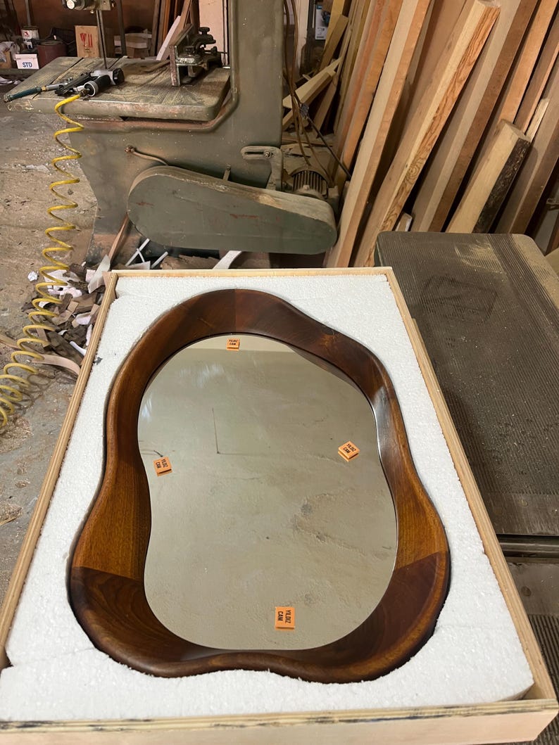 May include: An irregular, organic-shaped mirror with a polished dark brown wooden frame. The mirror surface features three small orange square stickers with the text YILDIZ CAM printed on them, and it is securely packed in a white foam-lined wooden crate.