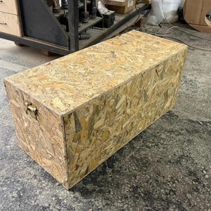 May include: A rectangular storage chest constructed from light brown oriented strand board, featuring a flat top surface and a single metallic handle attached to the side panel for lifting and carrying the box in a workshop environment.
