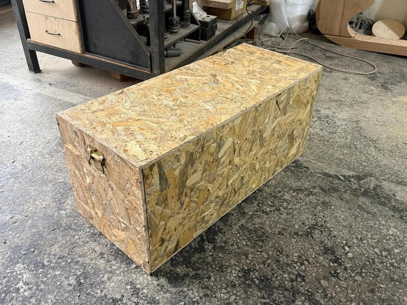 May include: A rectangular storage chest constructed from light brown oriented strand board, featuring a flat top surface and a single metallic handle attached to the side panel for lifting and carrying the box in a workshop environment.