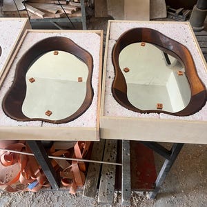 May include: Two irregular, organic-shaped mirrors with dark brown wooden frames, each nestled securely within a light-colored wooden shipping crate lined with protective white foam padding. Small square brown spacers are attached to the glass.