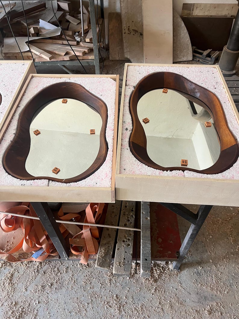 May include: Two irregular, organic-shaped mirrors with dark brown wooden frames, each nestled securely within a light-colored wooden shipping crate lined with protective white foam padding. Small square brown spacers are attached to the glass.