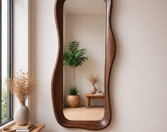 May include: A large, full-length mirror with a unique, wavy, dark brown wooden frame. The mirror is rectangular with softly curved edges. The reflection shows a room with plants and a wooden table. The mirror is mounted on a light-colored wall.