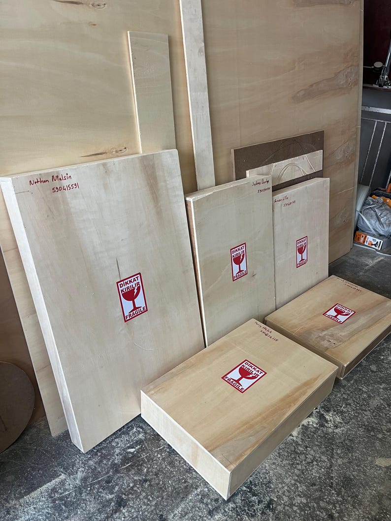 May include: Several rectangular light-colored wooden panels and boxes of varying sizes. Each piece features a white sticker with a red border and a red wine glass icon, containing the text DIKKAT KIRILIR and FRAGILE in bold black lettering.