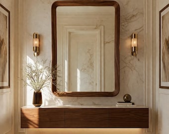 May include: A modern interior featuring a large, rectangular wooden mirror with rounded corners. Below the mirror is a floating wooden cabinet with three drawers. The wall is marble with two sconces and two framed pictures. A vase with branches sits on the cabinet.