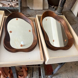 May include: Two irregular, organic-shaped wall mirrors with dark brown wooden frames. Each mirror is nestled in a light-colored wooden crate lined with white foam. The reflective surfaces feature small, square, orange adhesive tabs and one white bulb.