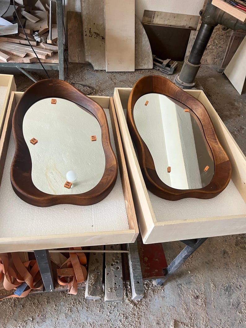 May include: Two irregular, organic-shaped wall mirrors with dark brown wooden frames. Each mirror is nestled in a light-colored wooden crate lined with white foam. The reflective surfaces feature small, square, orange adhesive tabs and one white bulb.
